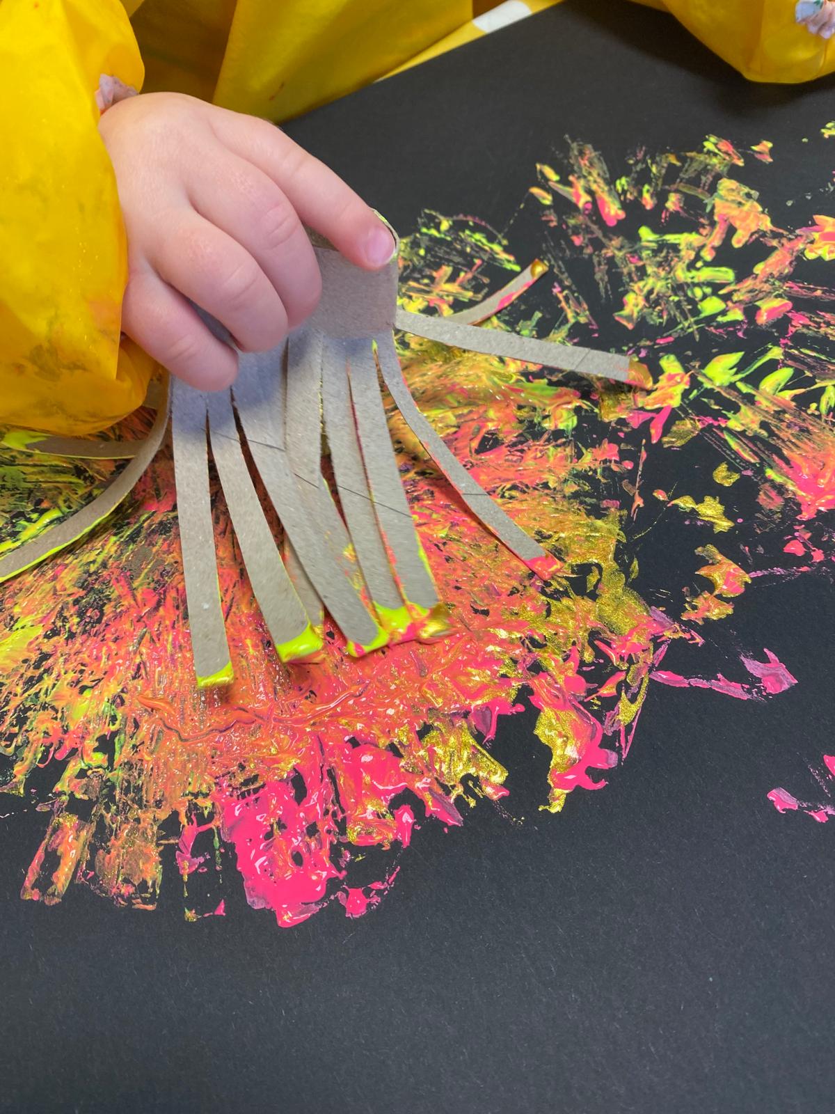Eine Kinderhand druckt mit einer eingeschnittenen Papprolle Farbe auf schwarzes Papier