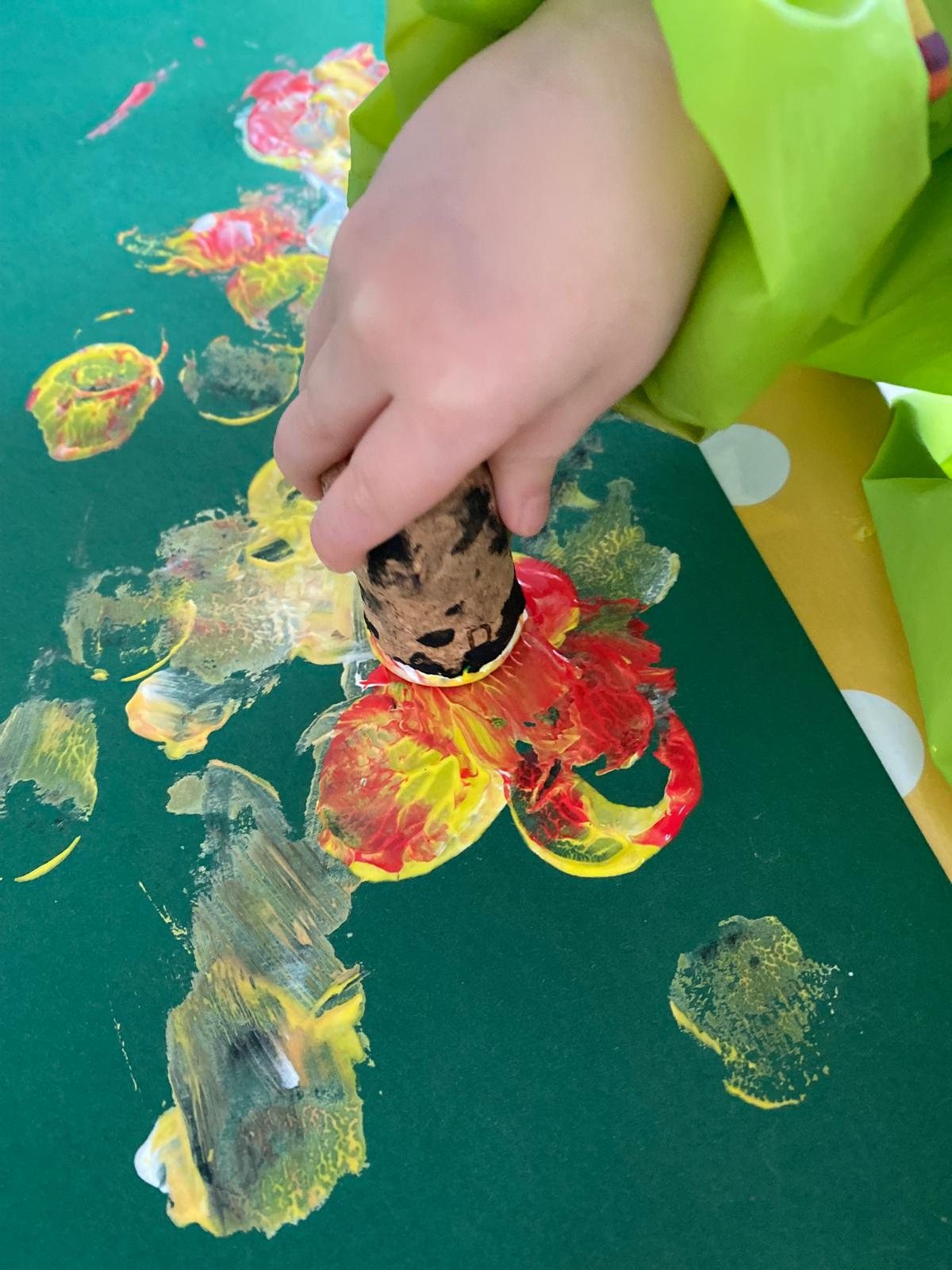 Eine Kinderhand stempelt mit einem Korken bunte Farbe auf grünes Papier